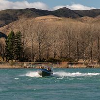 A photo of a jetboat taken from the far riverbank. In the background is a row of leafless trees and a series of hills