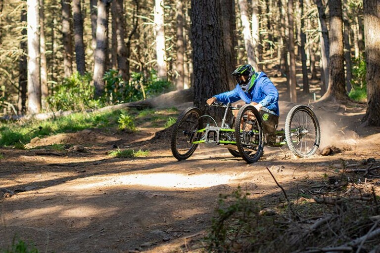 A photo of a person using the gravity quad bike. It is taken in a lightly wooded area from the side of the track just before they come past. They are wearing a full-face helmet and a blue hoodie