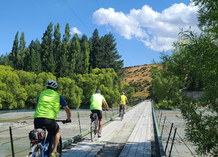 A photo of three people cycling over a large suspension bridge. The weather is clear