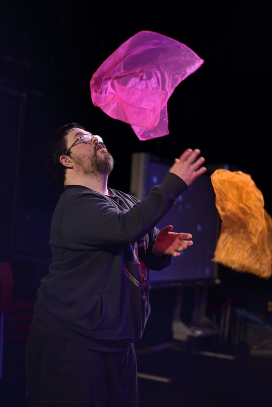 A photo of a bearded man with glasses looking a pink square of cloth that's been thrown in the air