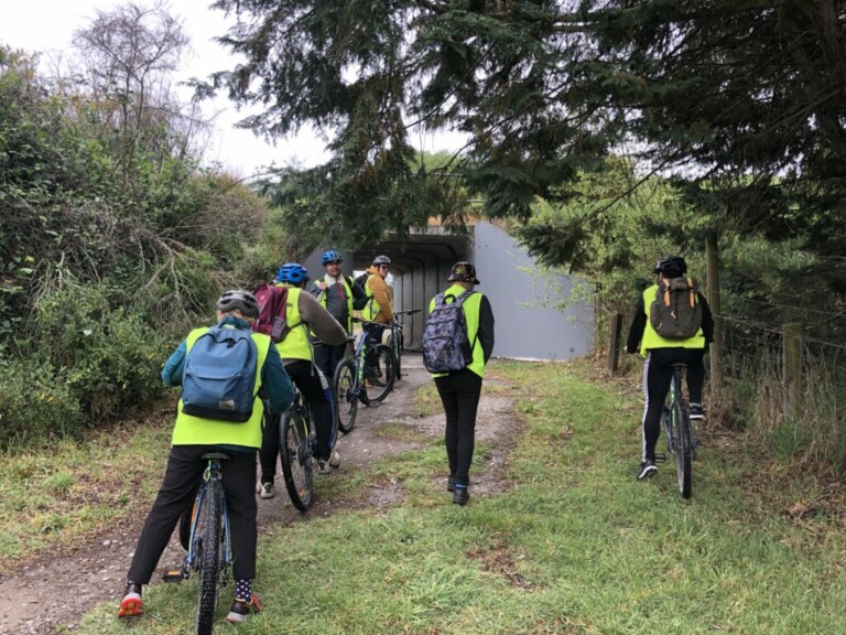 A photo of several people in single file on bicycles. One person walks to the right of them and another person on a bike is to the right of the walker. They are all wearing high visibility vests